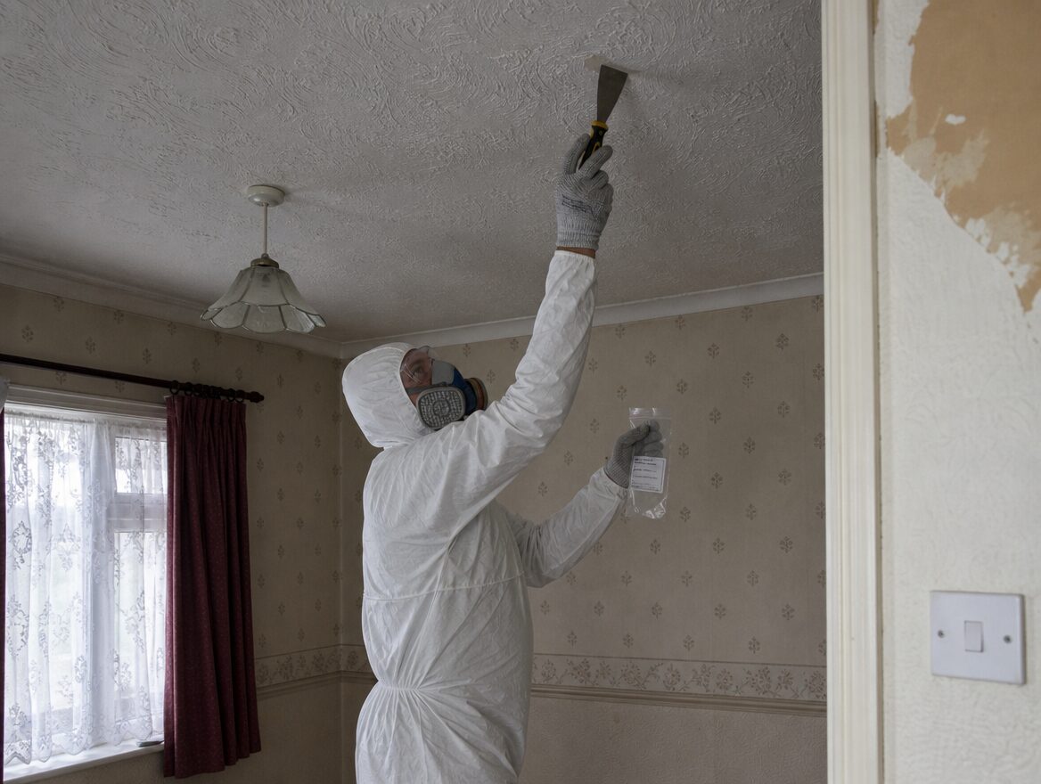 Surveyor in protective overalls taking a sample from an artex ceiling